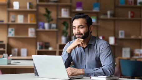Smiling bearded indian businessman working on laptop in modern office lobby space. Young indian student using computer remote studying, watching online webinar, virtual training on video call meeting.