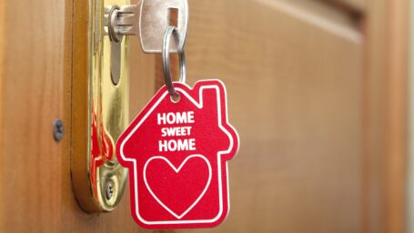 A key to a home in the door with a red key chain the shape of a house that says Home Sweet Home